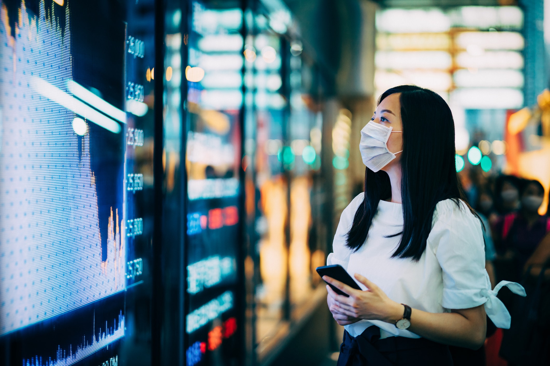 A woman with a mask looking at huge screens with market data