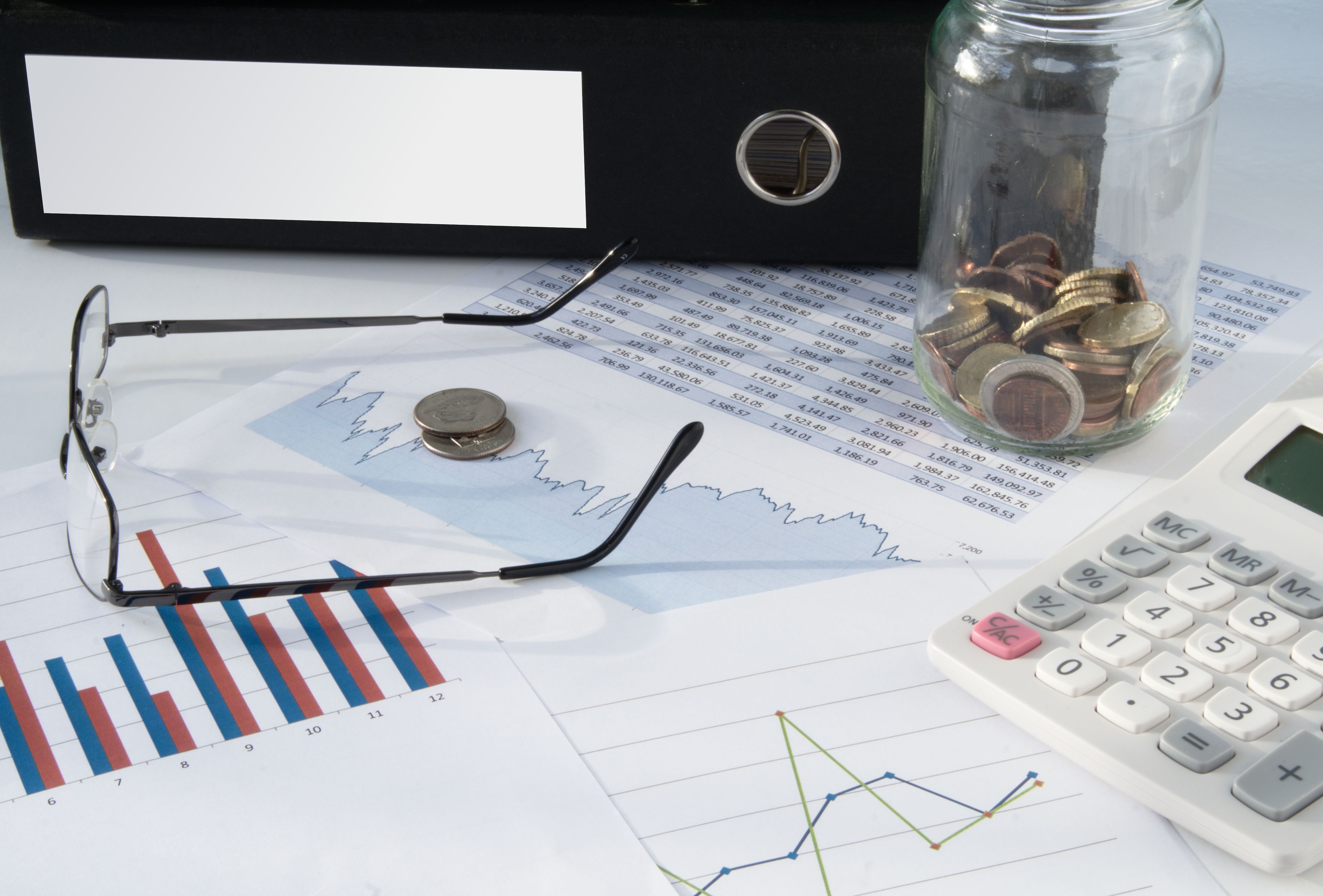 A shot of a desk covered in various items: a pair of glasses, a jar of coins, a binder, a calculator and several charts.