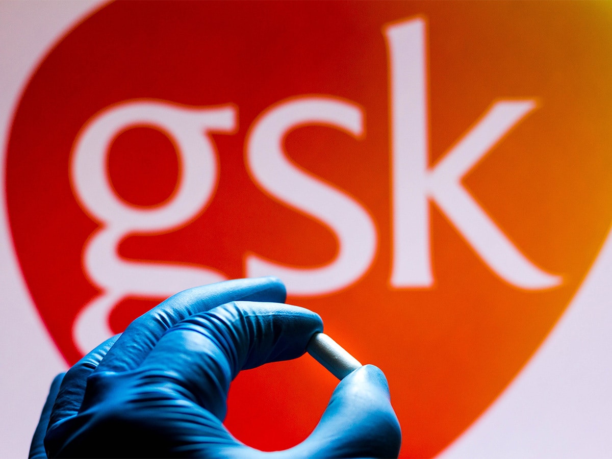 A blue gloved hand holding a pill in front of a GSK logo